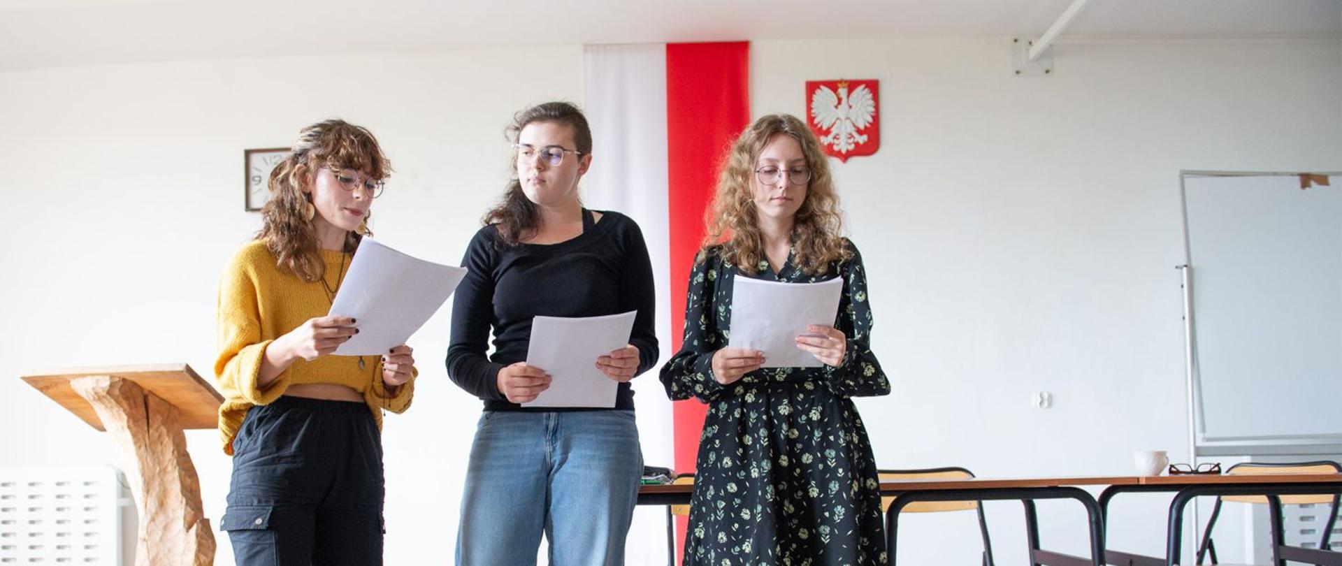 Trójka uczennic czytająca z kartek Ballady i romanse Adama Mickiewicza. W tle na białej ścianie flaga Polski oraz obok zawieszone godło Polski.