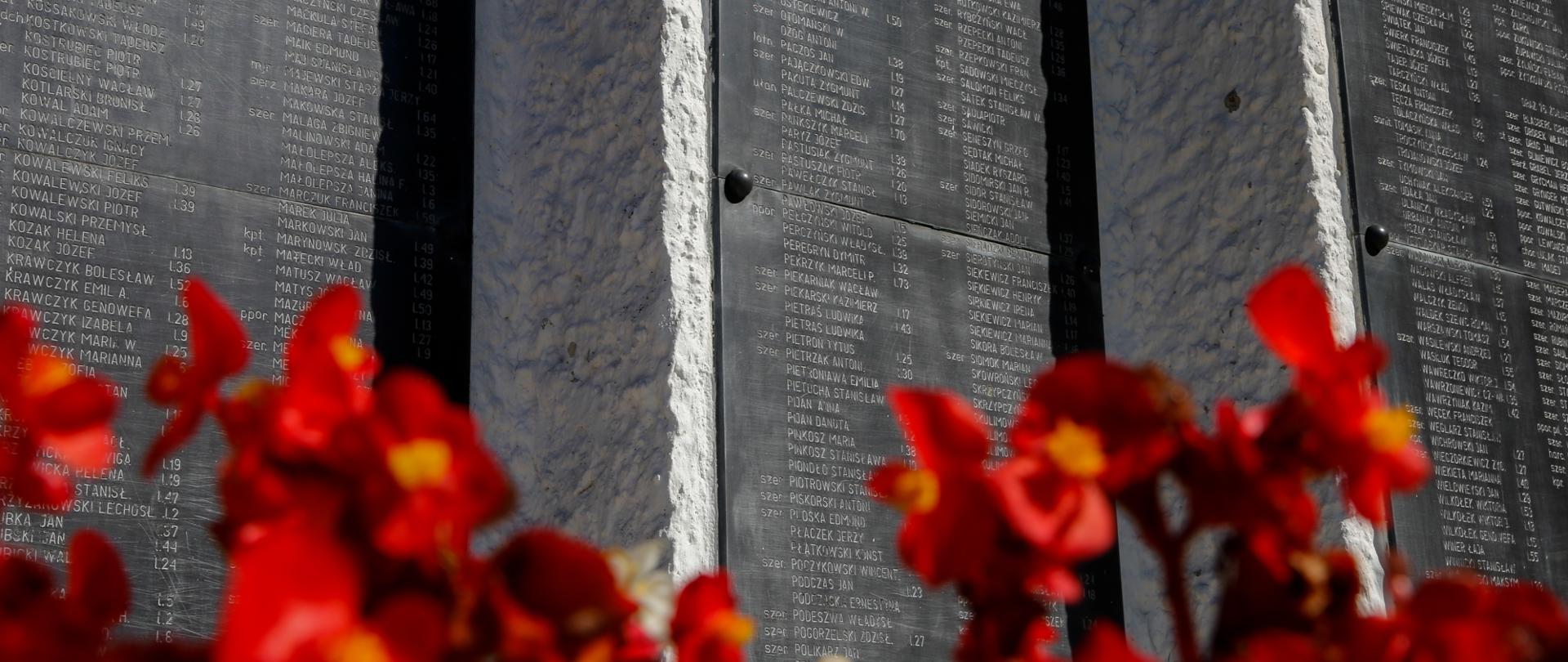 Tablice pomnika z nazwiskami osób zamordowanych. Na pierwszym planie kwiatki.