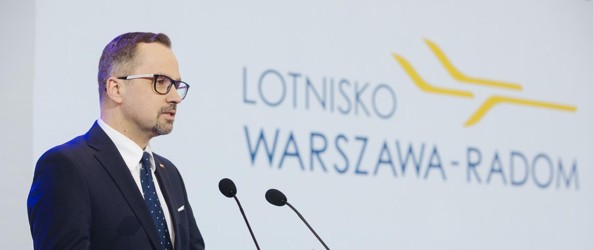Wiceminister Marcin Horała stoi przed pulpitem i mówi do dwóch mikrofonów.