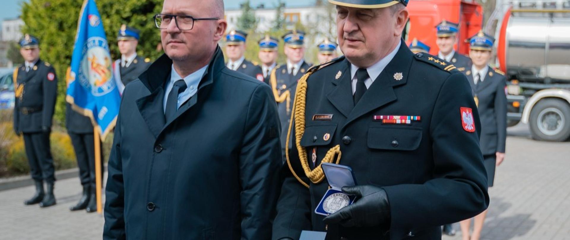 Prezydent - mężczyzna w granatowym płaszczu i Komendant w mundurze galowym prezentują medal. W tle załogo w mundurach galowych.