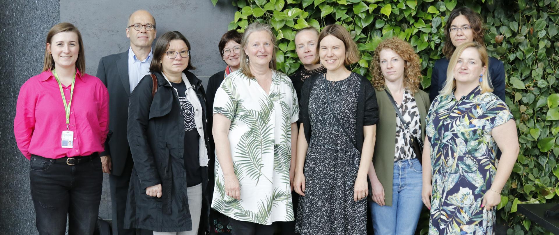 Zdjęcie grupowe. Dziewięć uśmiechniętych kobiet i jeden mężczyzna stoją na tle ścianki utworzonej z zielonych roślin, 