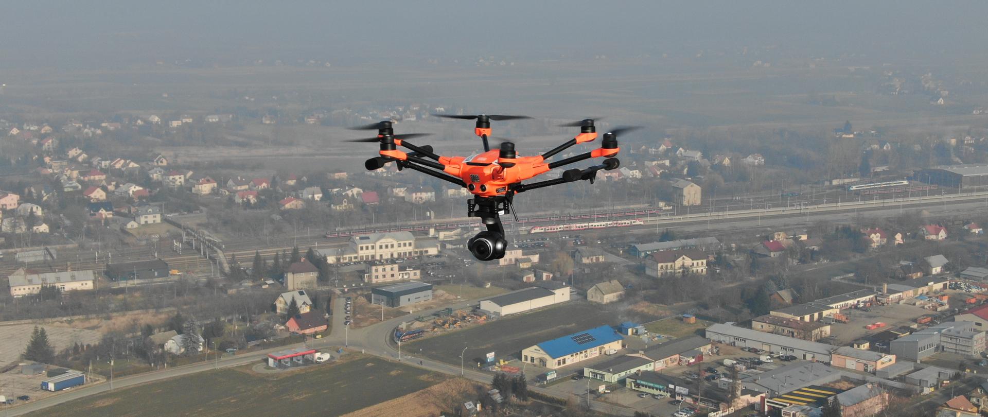 Zdjęcie robione w powietrzu przedstawia sześciowirnikowego lecącego drona z kamerą w kształcie kuli podwieszoną u dołu. W tle lekko zamglony kadr miasta z widoczną linią kolejową i dwoma pociągami osobowymi stojącymi na stacji.