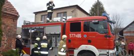 Pożar budynku mieszkalnego w Wydawach. Strażacy OSP Zielona Wieś po zakończonej akcji, wkładają sprzęt na półki samochodu ratowniczo-gaśniczego. Jeden z nich stoi na dachu wozu.W tle budynki.