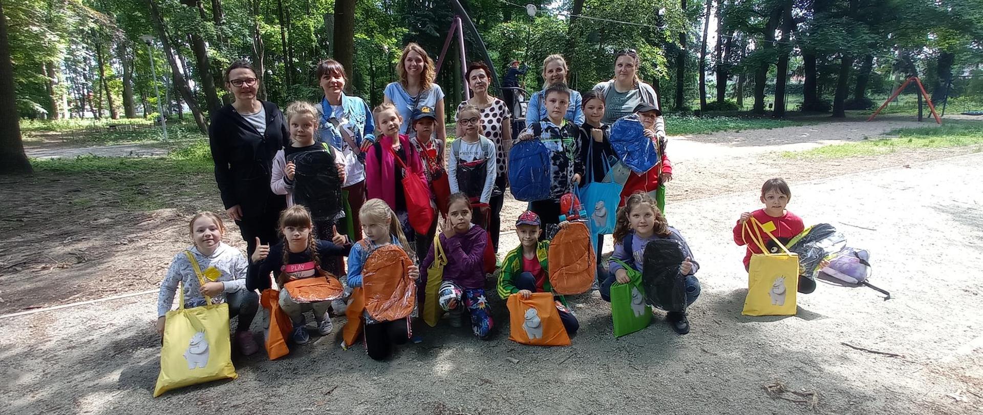 Grupa kilkanaściorga dzieci i sześć kobiet pozuje do zdjęcia w parku. Za ich plecami zielone drzewa. Dzieci trzymają w rękach kolorowe torby i plecaki. 