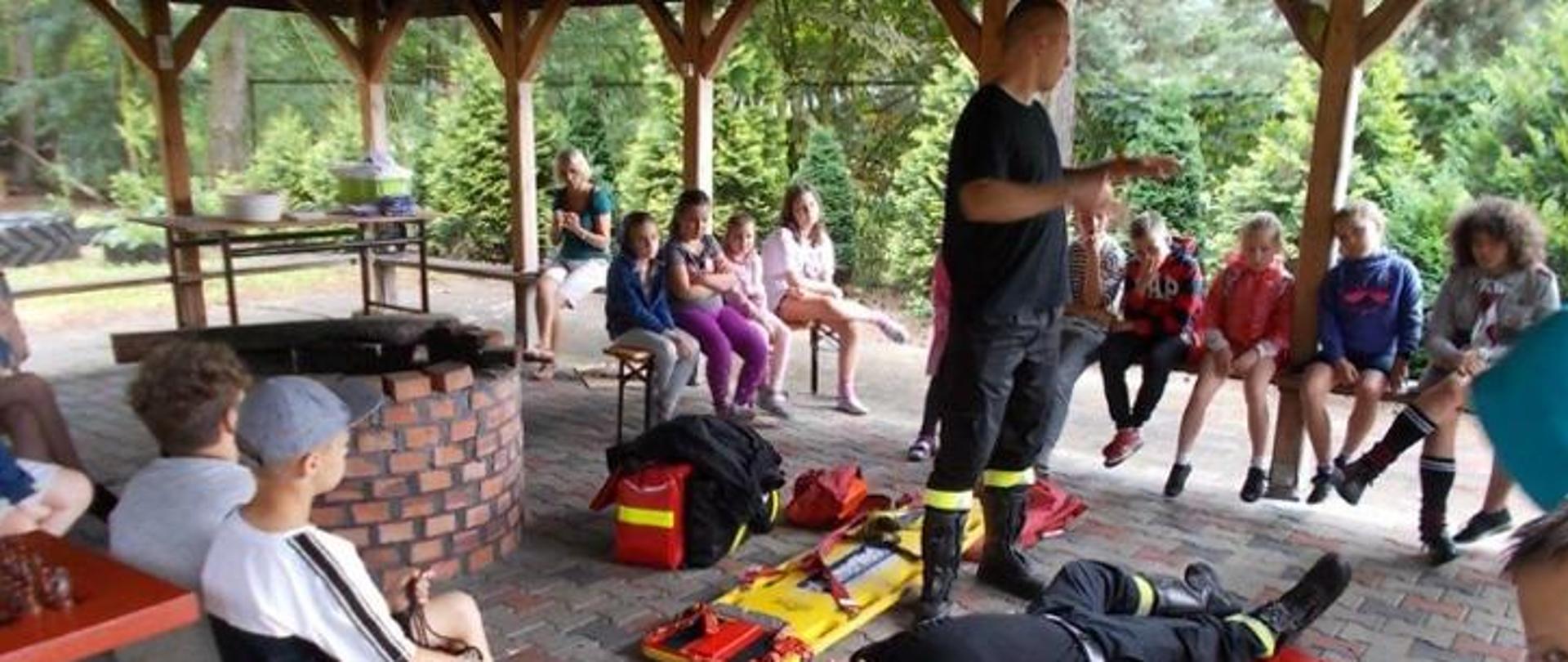 strażak prezentuje dookoła zebranym dzieciom zasady pierwszej pomocy przedmedycznej 