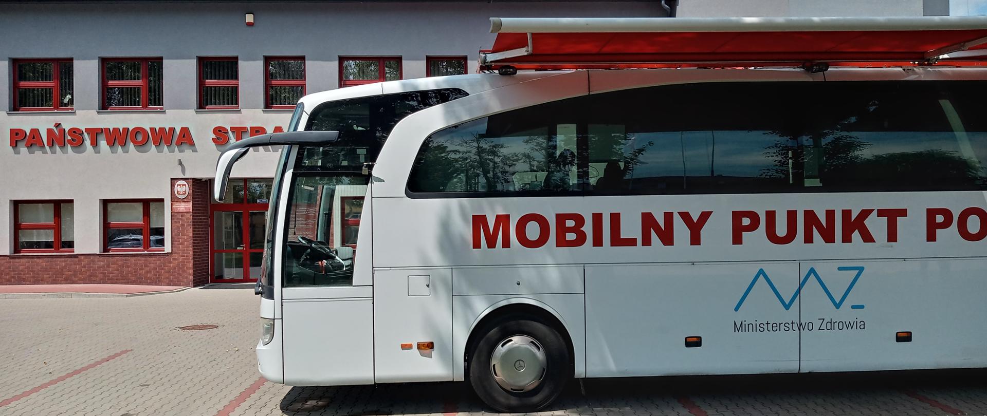Przed szarym, dwukondygnacyjnym budynkiem Państwowej Straży Pożarnej stoi biały autobus, na którym widnieje napis: mobilny punkt poboru krwi.