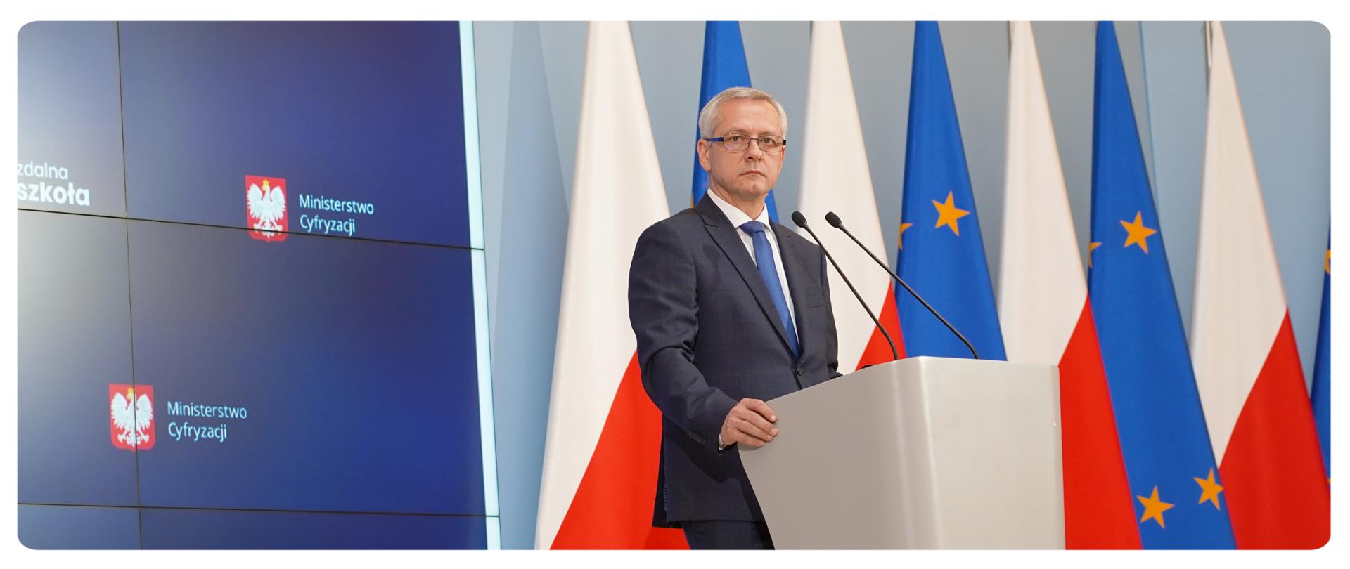 Minister Marek Zagórski przy mównicy, patrzy w prawą stronę. Za nim na ekranie fragment prezentacji - widać dwa logotypy MC na granatowym tle. Po lewej stronie ministra flagi: polskie i unijne
