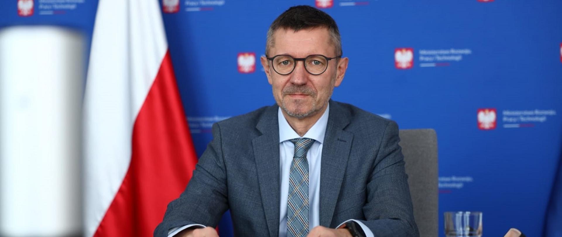 Wiceminister rozwoju, pracy i technologii Robert Tomanek siedzący przy stole, z tyłu za jego plecami flaga Polski i baner MRPiT