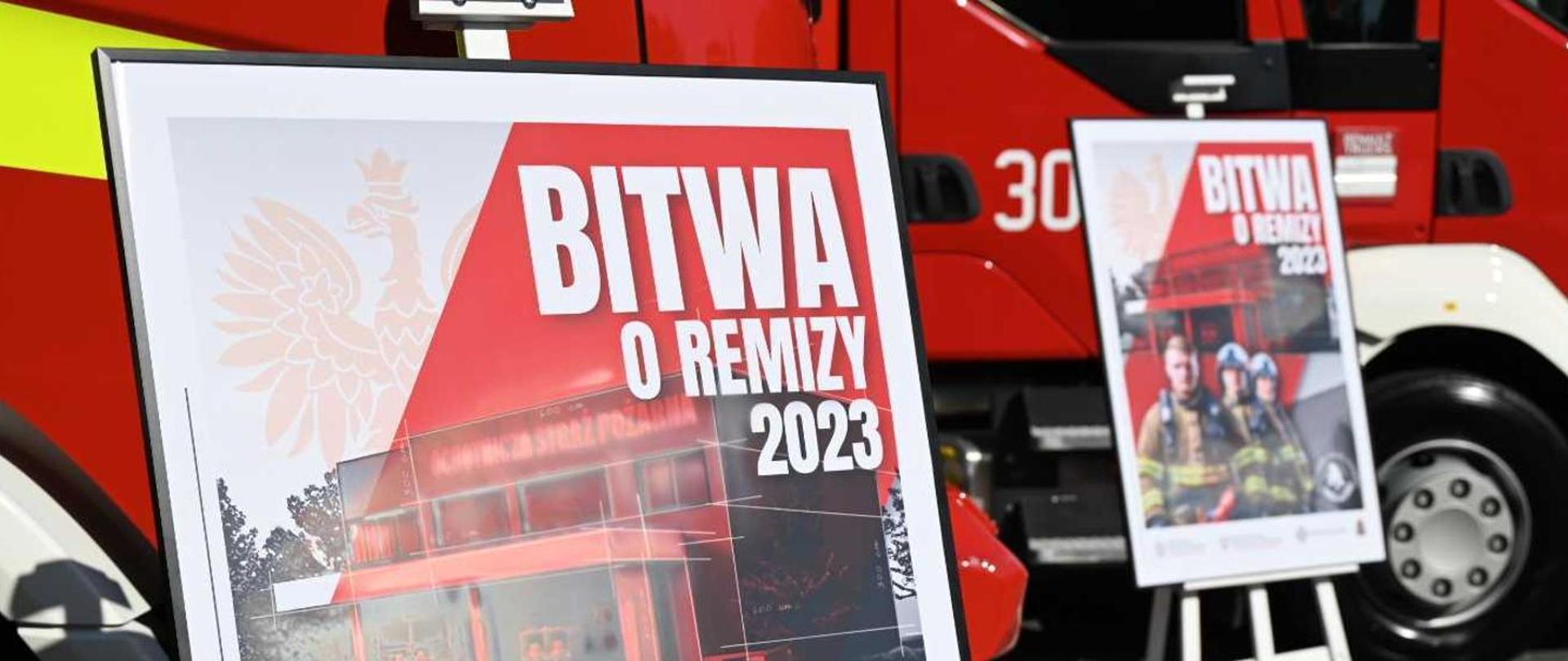Na tle samochodu wozu straży pożarnej widac dwa plakaty promujące akcję "Bitwa o Remizy 2023".