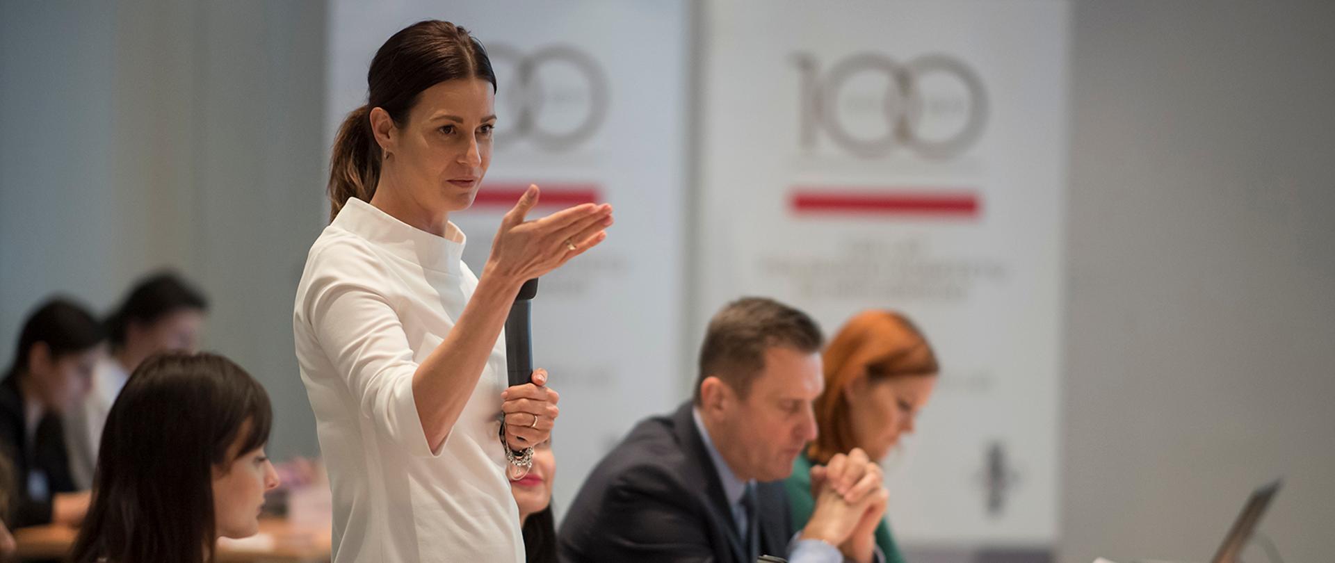 Minister Sportu Danuta Dmowska-Andrzejuk stoi przy stole prezydialnym i przemawia. W tle rozmyte logo 100-lecia Polskiego Komitetu Olimpijskiego