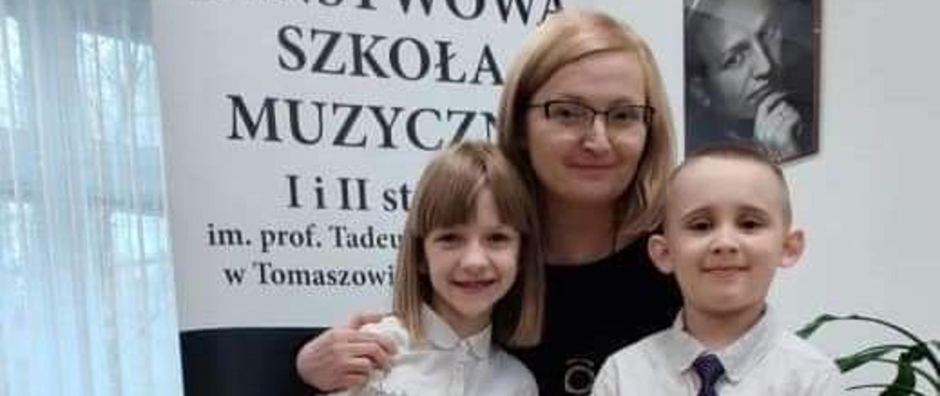 Dziewczynka i chłopiec wraz z nauczycielką na tle roll- upa PSM w Tomaszowie Mazowieckim