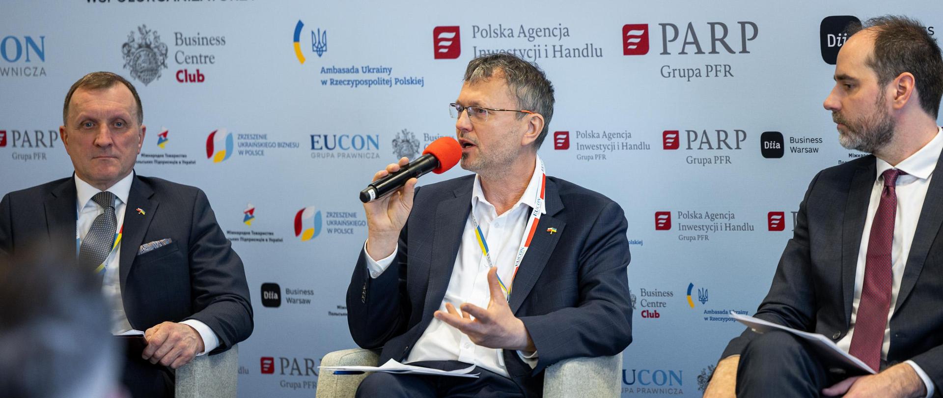 Wiceminister Jacek Bartmiński podczas panelu siedzi na kremowym fotelu z mikrofonem w ręku. Po prawej i lewej stronie siedzą dwaj inni paneliści. W tle ścianka z logotypami współorganizatorów, patronów i partnerów