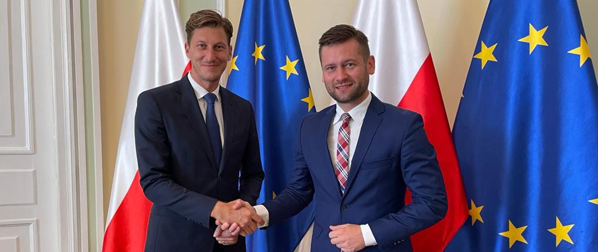 Minister Kamil Bortniczuk spotkał się z Prezydentem czeskiej Narodowej Agencji Sportowej. Minister i Prezydent czeskiej Narodowej Agencji Sportowej stoją na pierwszym planie, w tle flaga Polski i Unii Europejskiej.