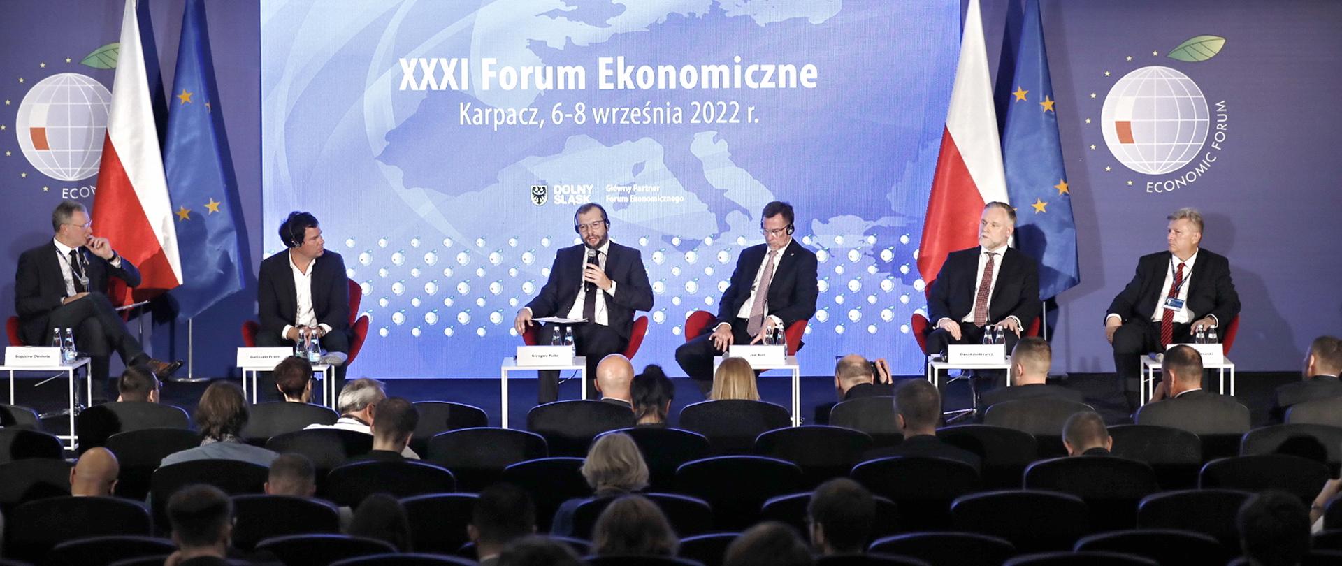  Minister Grzegorz Puda siedzi w fotelu środku na scenie, trzyma mikrofon w lewej ręce i mówi do niego. Obok niego siedzi pięć innych osób.