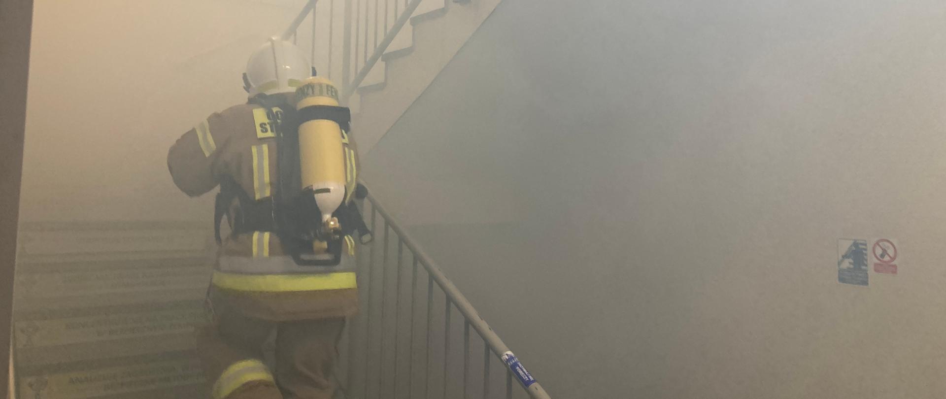Strażak ubrany w aparat ochrony dróg oddechowych wchodzi po schodach. W tle panuje zadymienie.