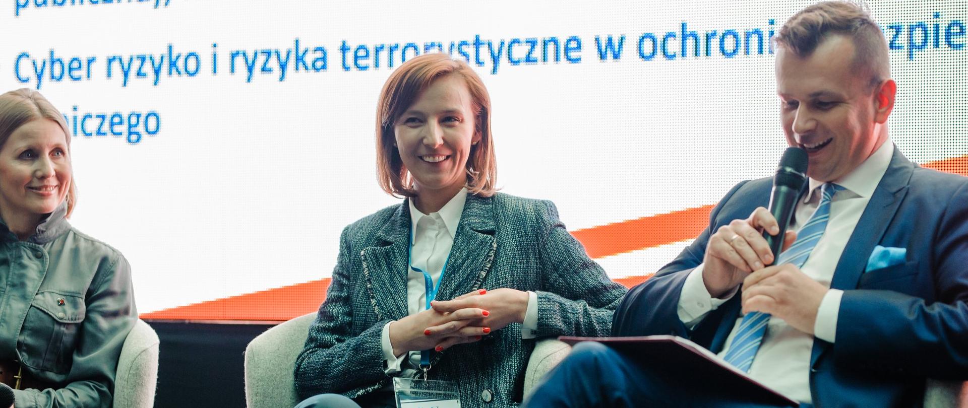Z prawej strony na fotelu siedzi wiceminister Adam Hamryszczak. Trzyma mikrofon przy ustach. Obok niego siedzą dwie kobiety.