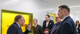 Minister Czarnek stoi w towarzystwie kilku mężczyzn w białym pomieszczeniu z oknem z żółtą szybą.