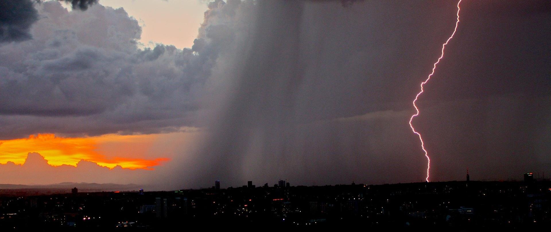 Zdjęcie przedstawia błyskawicę nad miastem i smugi podającego deszczu.