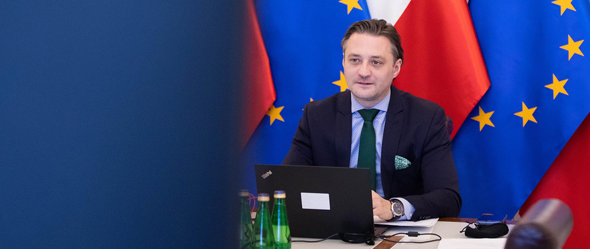 Wiceminister Bartosz Grodecki uczestniczący w Forum Schengen za pośrednictwem wideokonferencji na tle flag Polski i Unii Europejskiej.