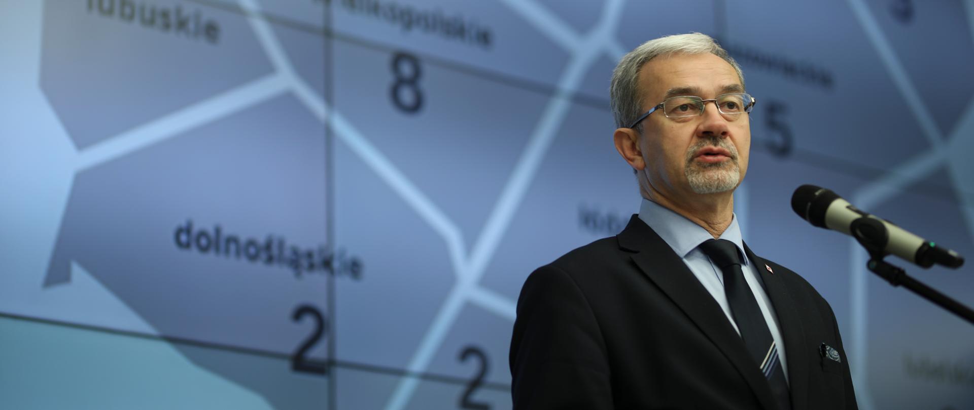 Minister Kwieciński mówi do stojącego mikrofonu na tle ekranu z wyświetloną mapą Polski