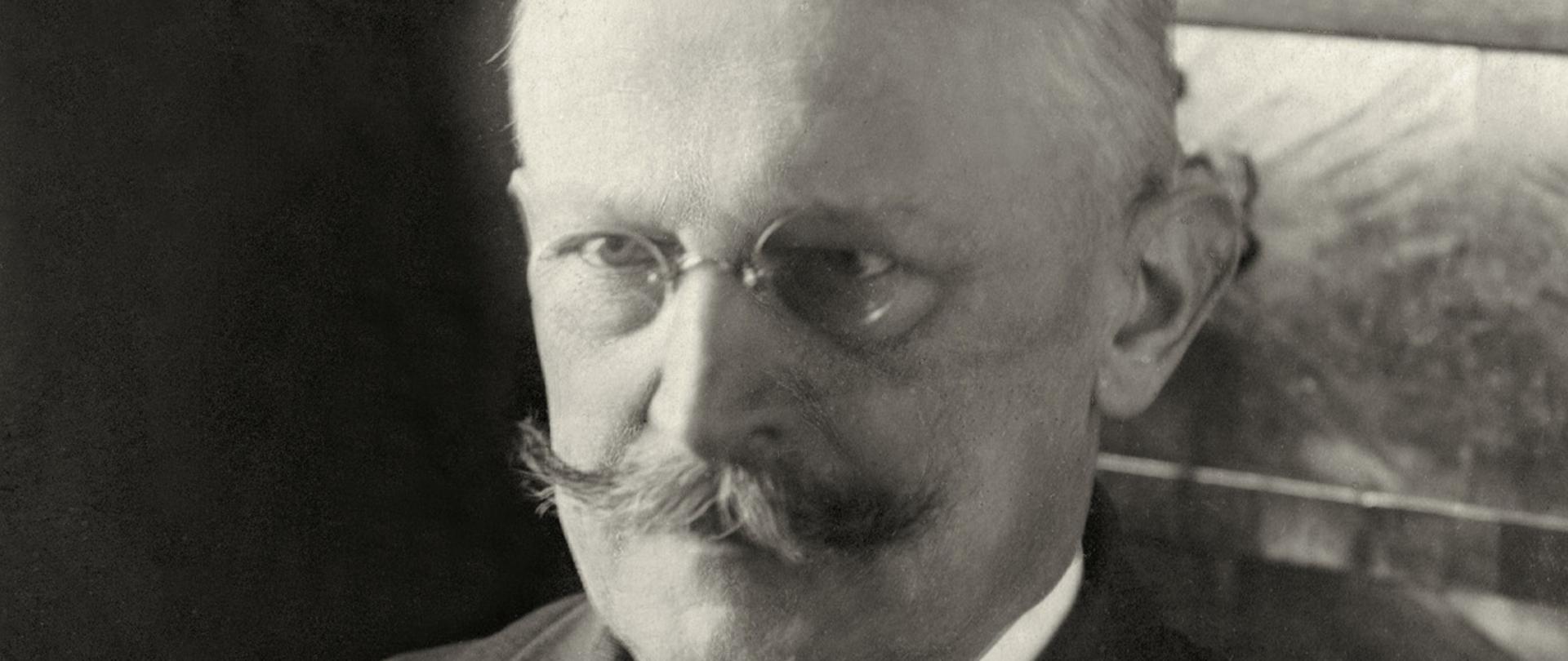 Stanisław Patek - minister spraw zagranicznych. Czarno-białe zdjęcie mężczyzny w okularach i wąsami, w ciemnej dwurzędowej marynarce, ciemny krawat.