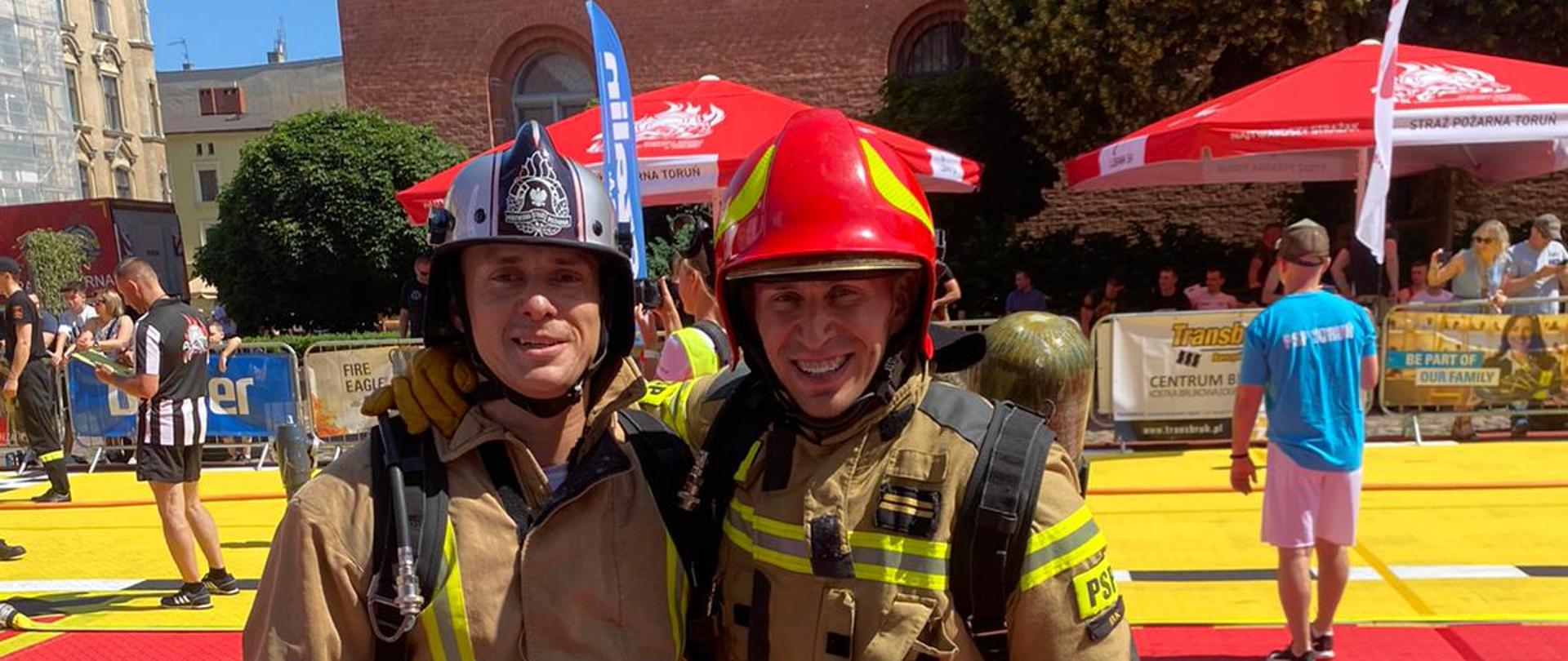 Zdjęcie przedstawia dwóch strażaków pozujących do zdjęcia na czerwonym dywanie ubranych w ubrania specjalne z hełmami na głowach.