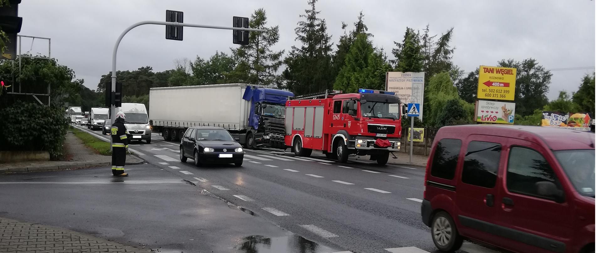 na skrzyżowaniu strażak kieruje ruchem na poboczu stoi rozbita ciężarówka i pojazd ratowniczo - gaśniczy 