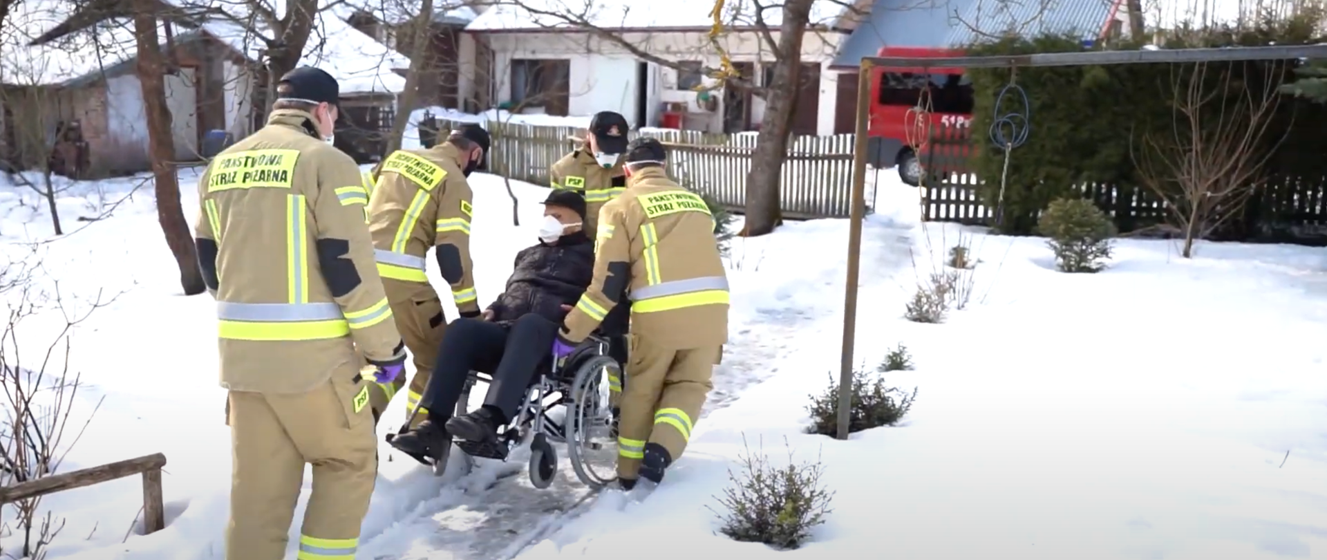 Strażacy pomagają osobie niepełnosprawnej transportując ją na wózku inwalidzkim przez zaśnieżoną ścieżkę do stojącego za bramką samochodu strażackiego
