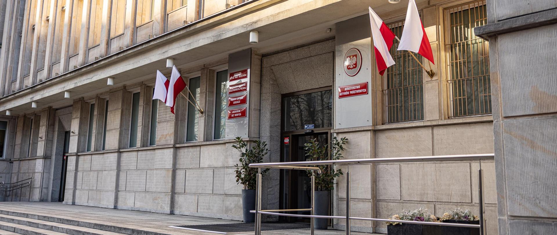 Budynek Ministerstwa Aktywów Państwowych. Na elewacji wiszą tabliczki oraz flagi biało-czerwone. Przed wejściem kwiaty. 
