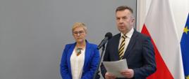 Minister Wieczorek i wiceminister Mrówczyńska stoją na sali, minister trzyma w ręku kartki papieru i mówi do mikrofonu na stojaku, za nimi pod szarą ścianą flagi Polski i UE.
