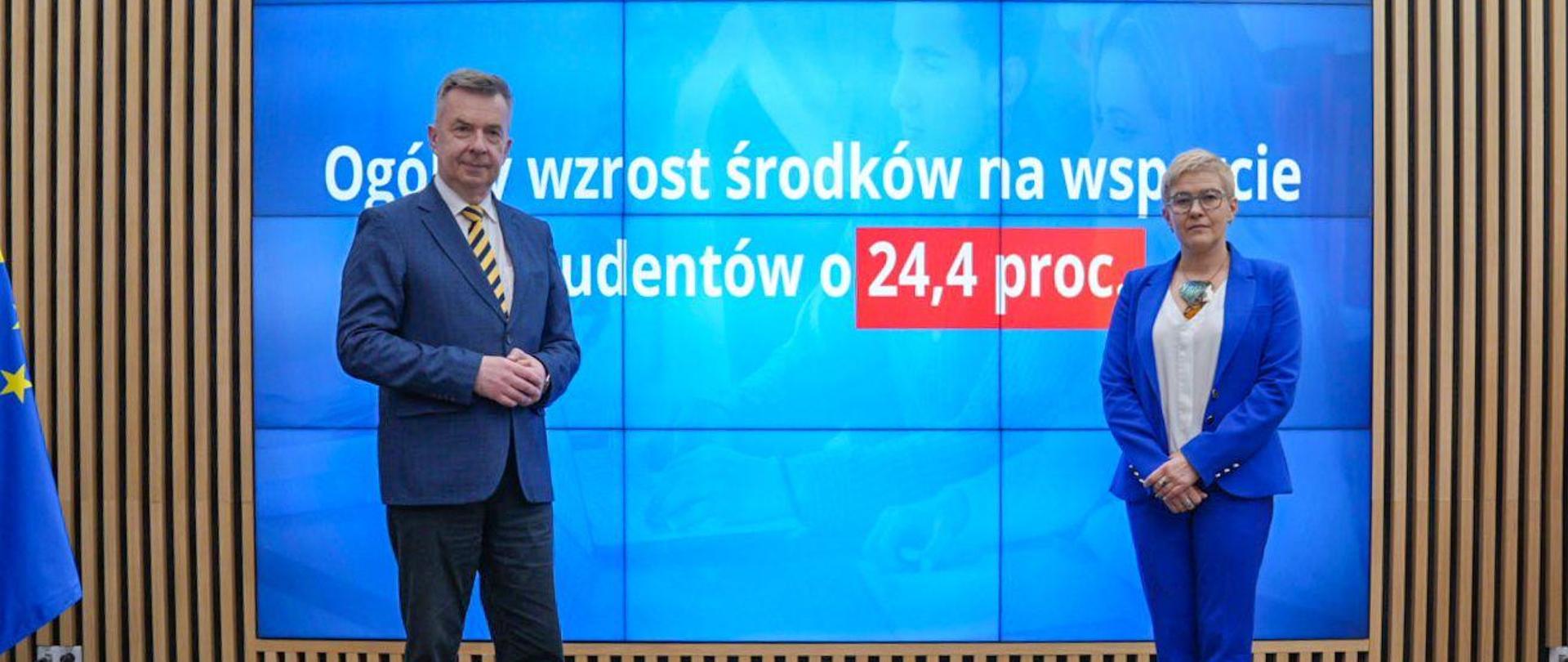 Minister Wieczorek i wiceminister Mrówczyńska stoją na sali, za nimi na wbudowanym w ścianę wielkim ekranie napis Ogólny wzrost środków na wsparcie studentów o 24,4 proc.