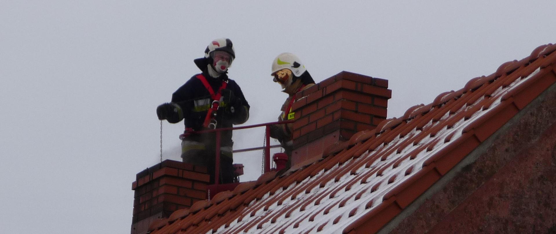 Zdjęcie przedstawia dach budynku jednorodzinnego pokryty pomarańczową dachówką. na dachu znajdują się dwa kominy, jeden z nich pali się. Na dachu znajduje się dwóch strażaków, którzy czyszczą przewód kominowy.