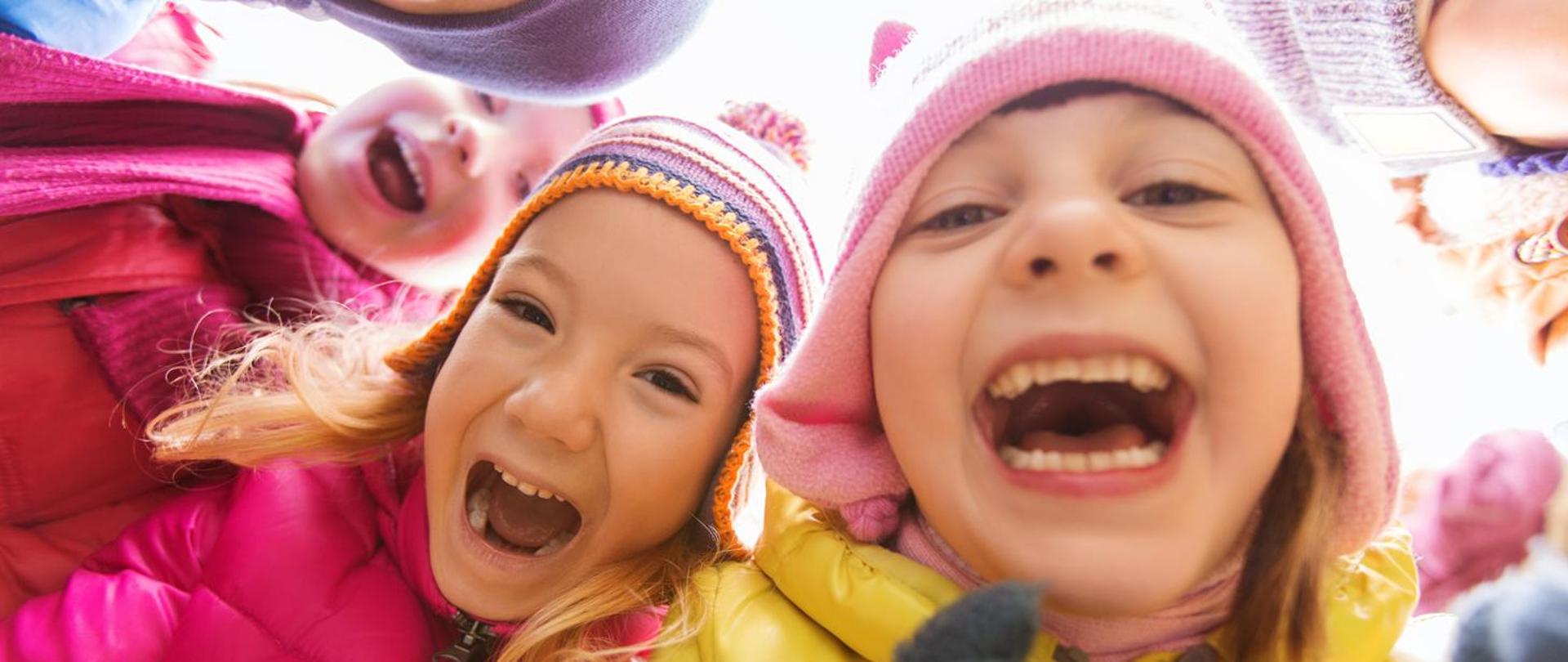 Zdjęcie przedstawia trójkę uśmiechniętych dzieci, ubranych w ciepłe kurtki i czapki.