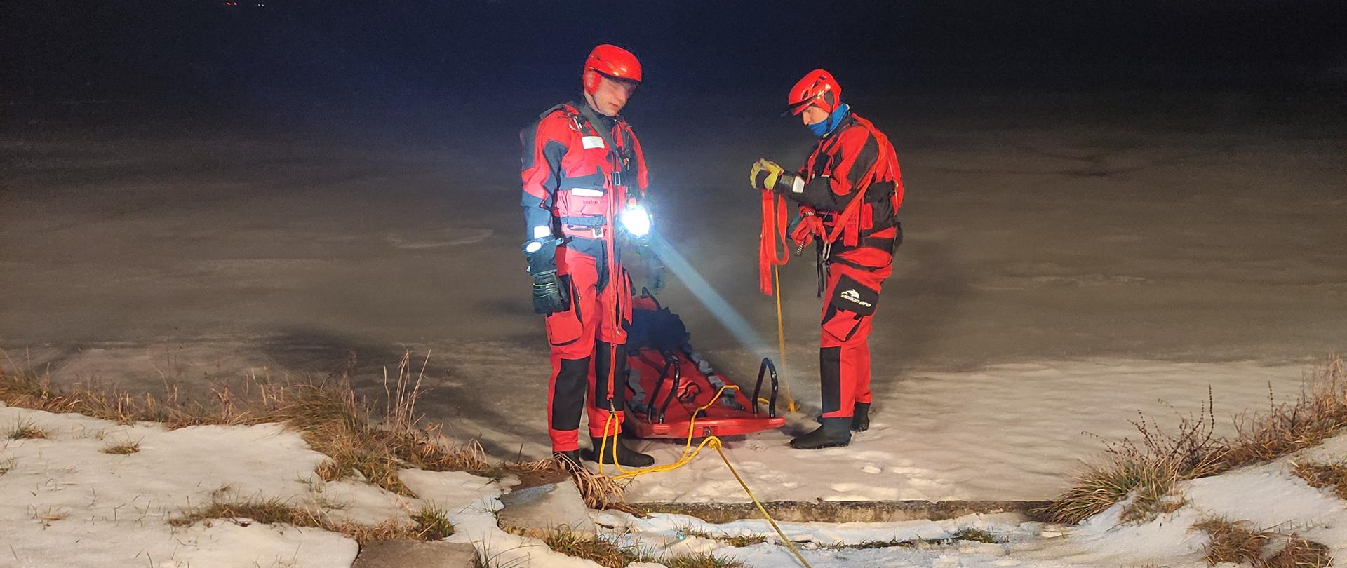 Dwóch strażaków w czerwonych skafandrach lodowych przygotowuje się do wejścia na zamarznięty staw, warunki nocne, jeden świeci latarką.