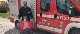 Zdjęcie przedstawia druha OSP w Dunajku z osobą starszą poruszającą się na wózku, na tle pojazdu pożarniczego, którym zostanie dowieziona do punktu szczepień