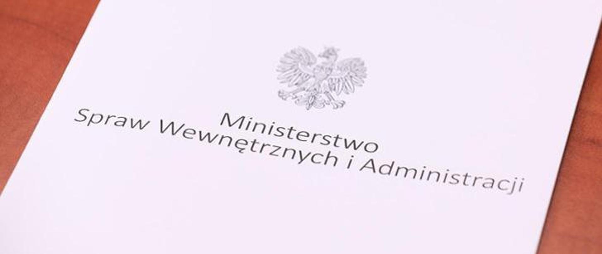 Biała teczka leżąca na stole, na którym widnieje napis Ministerstwo Spraw Wewnętrznych i Administracji i orzeł Polski.