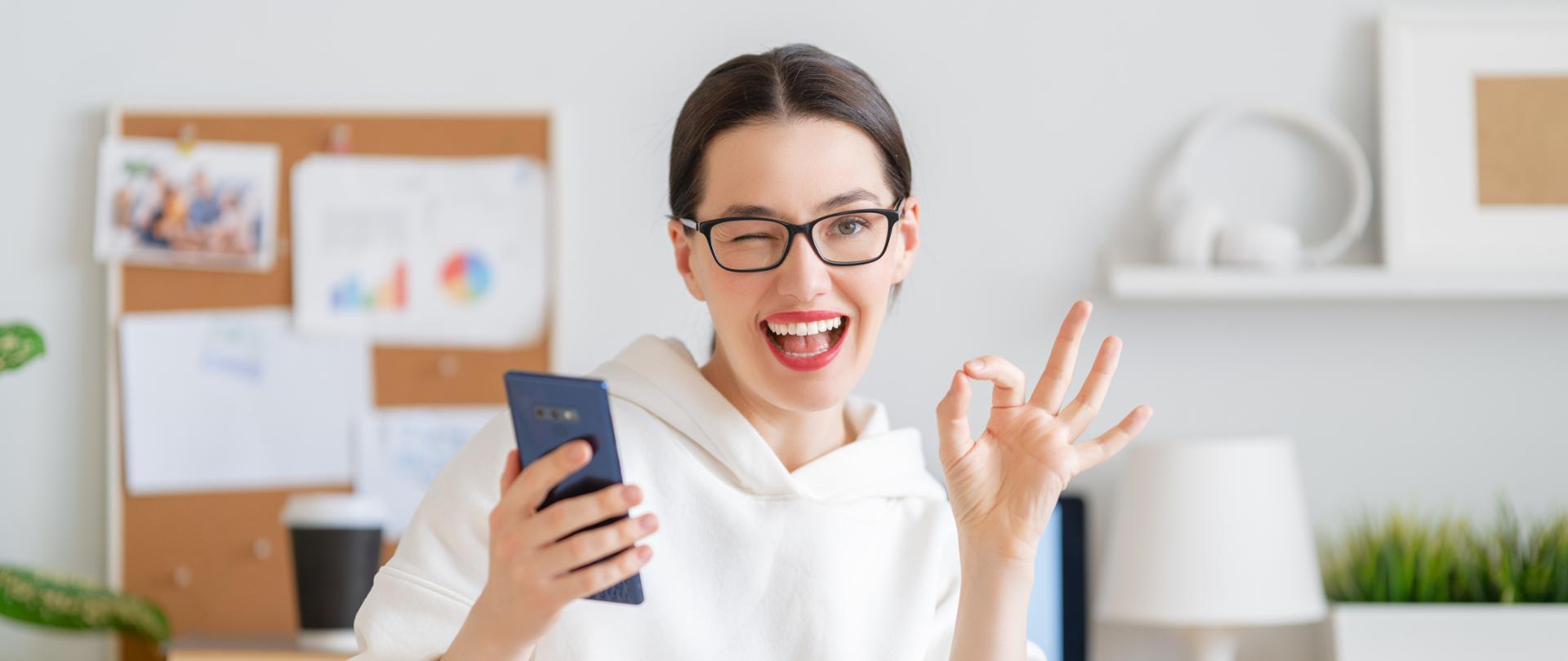 Uśmiechnięta kobieta trzymają w prawej dłoni smartfon, lewą pokazuje znak ok, puszcza oko.