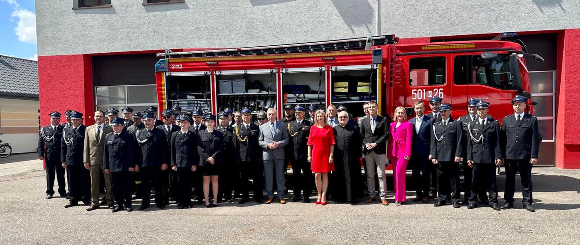 Zaproszeni goście oraz strażacy biorący udział w uroczystości stoją przed samochodem pożarniczym.