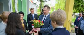Minister wręcza bukiet kwiatów nauczycielom, stoją przed wejściem do szkoły.
