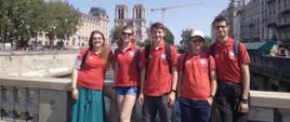 Grupa młodzieży w czerwonych koszulkach stoi na kamiennym moście, za nimi panorama miasta.