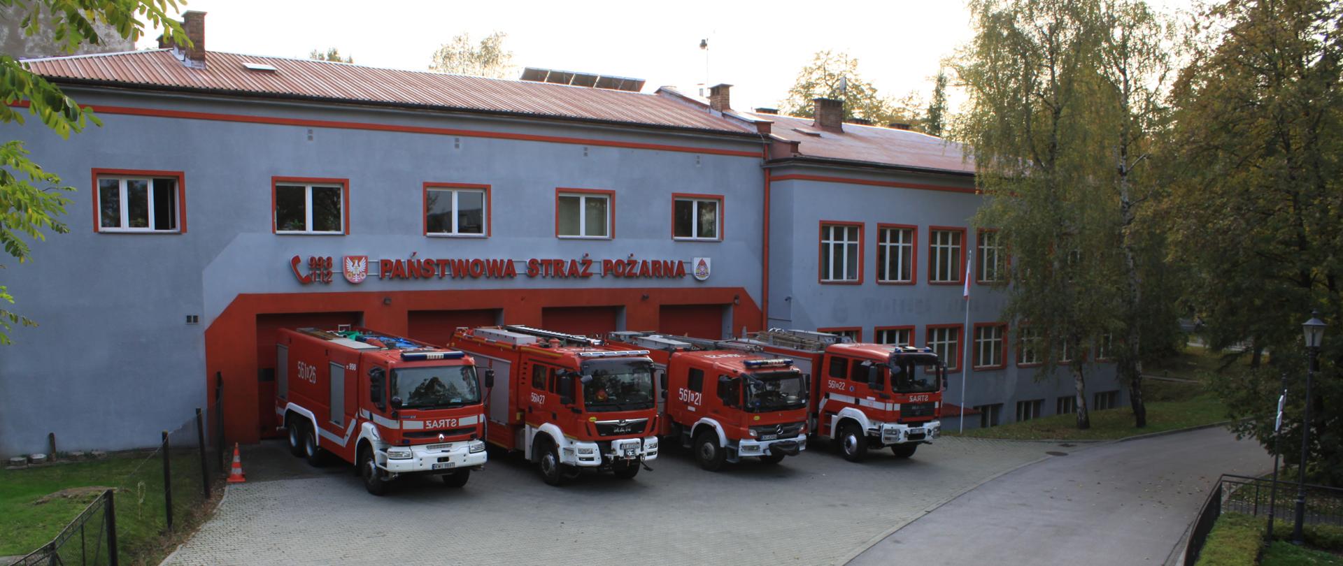 Zdjęcie zrobione na tle fasady budynku Komendy Powiatowej Państwowej Straży Pożarnej w Wieliczce od strony stawu na teranie Kopalni soli Wieliczka. Przed czterema bramami garażowymi stoją ciężarowe samochody strażackie. 