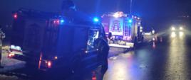 Dwa wozy strażackie stoją na poboczu drogi z włączonymi światłami błyskowymi oraz oświetleniem terenu akcji, jest noc, w oddali z przeciwka nadjeżdża samochód. 