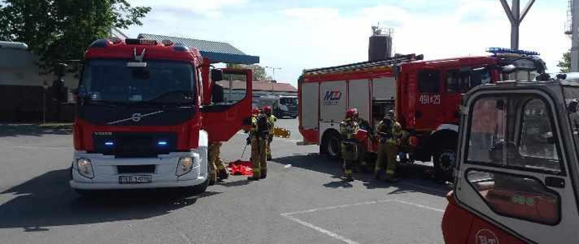 Zdjęcie przedstawia samochody pożarnicze stojące na placu manewrowym zakładu. Strażacy przygotowują się do wejścia strefę zagrożenia. 