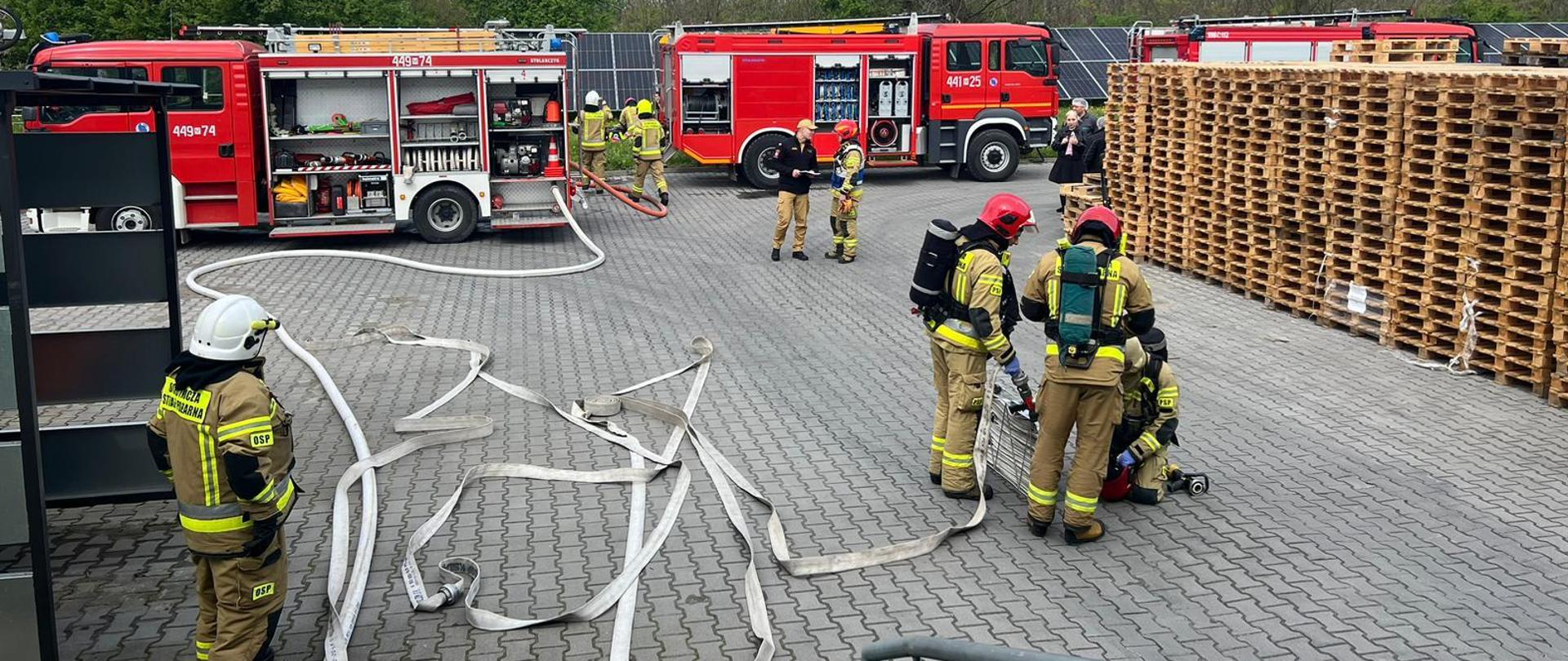 Strażacy przy samochodach pożarniczych, z których rozwinięte są linie gaśnicze troje ze strażaków przygotowuje się do wejścia do budynku szykują sprzęt zakładają aparaty powietrzne 