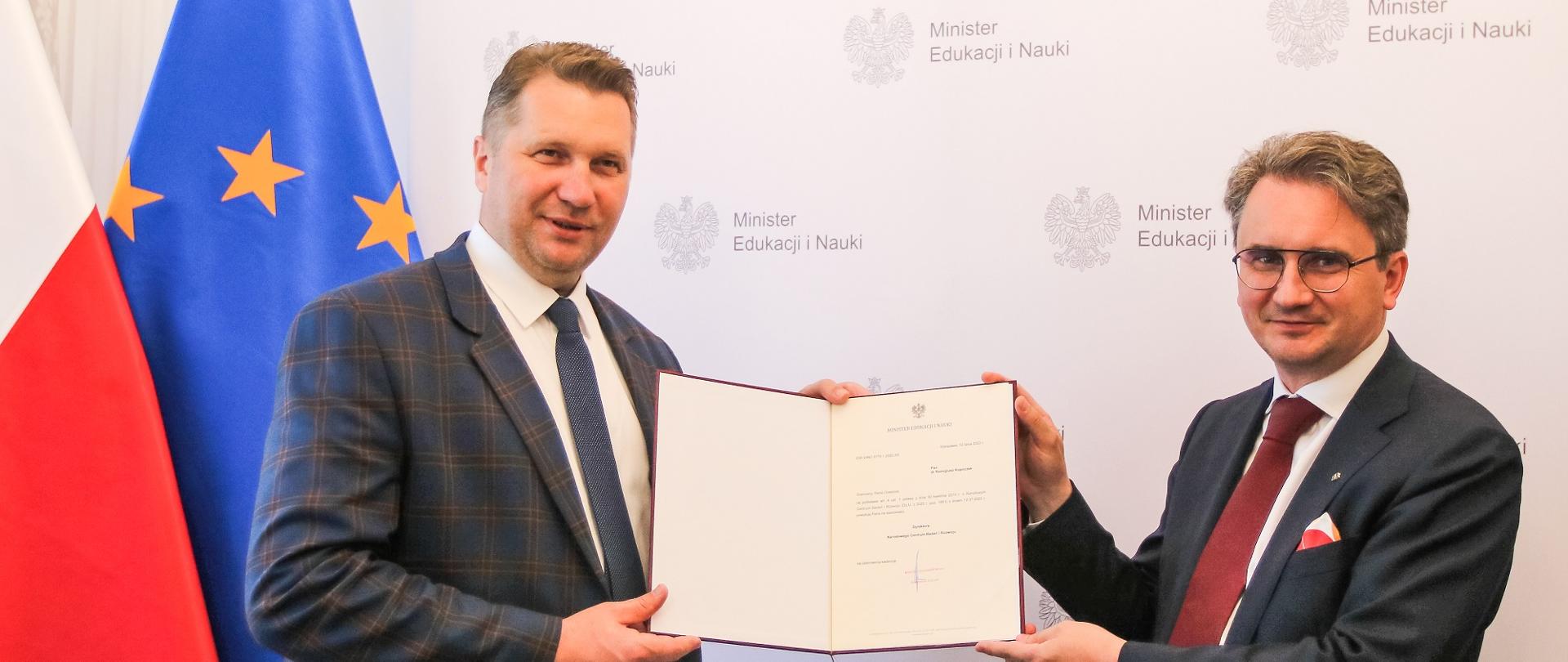 Minister Czarnek wręcza nowemu dyrektorowi NCBR otwarty dokument, za nim flagi Polski i UE.
