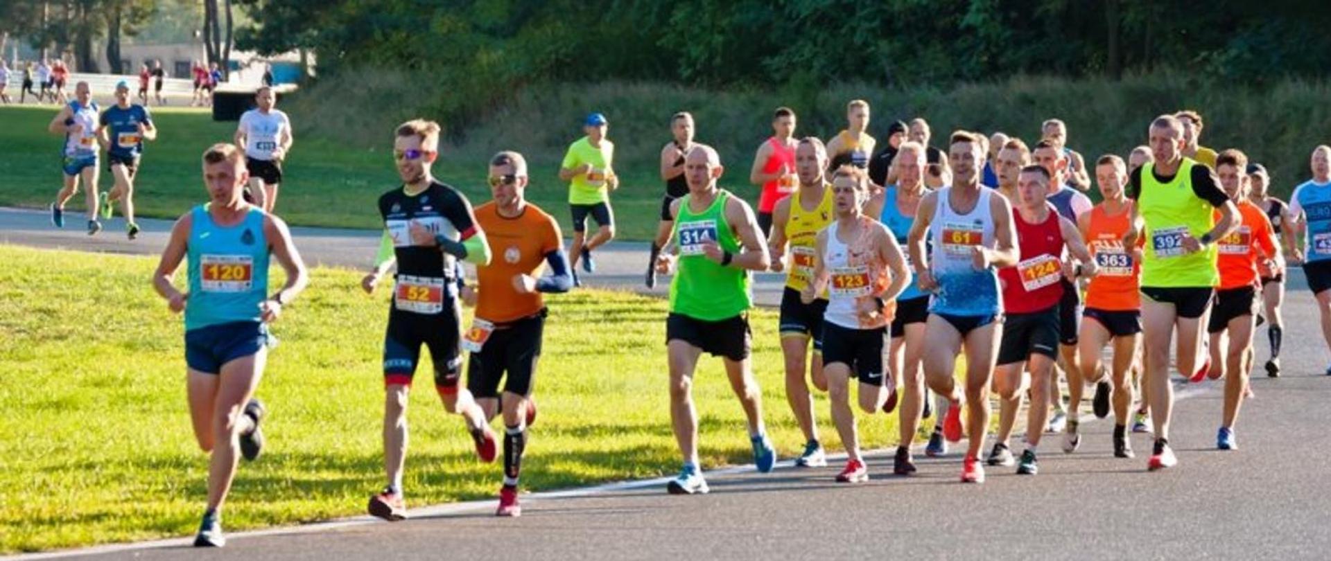 Zdjęcie przedstawia ciąg biegaczy podczas letnich zawodów sportowych. 