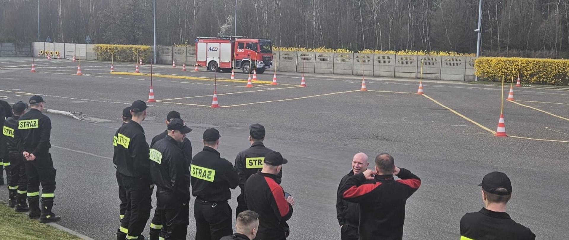 Na zdjęciu strażacy stojący przy placu manewrowym, na którym jeden z kierowców w samochodzie ratowniczo-gaśniczym wjeżdża w wyznaczone pachołkami ciasne miejsca tak aby nie potrącić żadnego z nich.