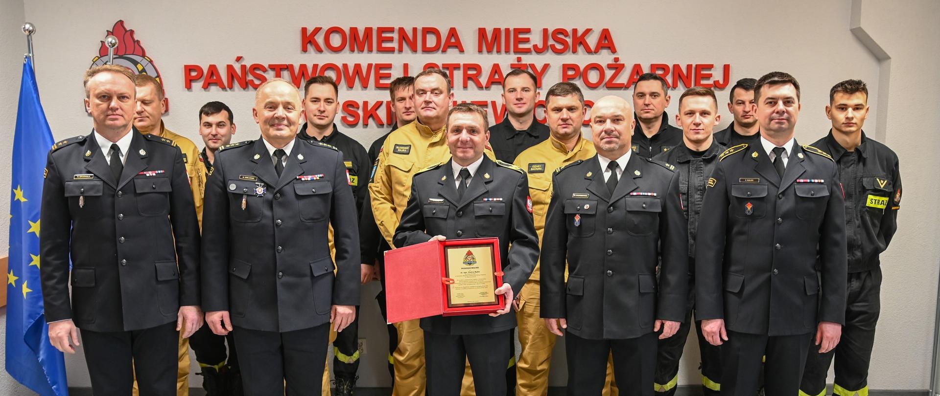 Strażacy w mundurach wyjściowych, służbowych i ubraniach koszarowych stoją na tle białej ściany z czerwonym napisem Komenda Miejska Państwowej Straży Pożarnej w Skierniewicach