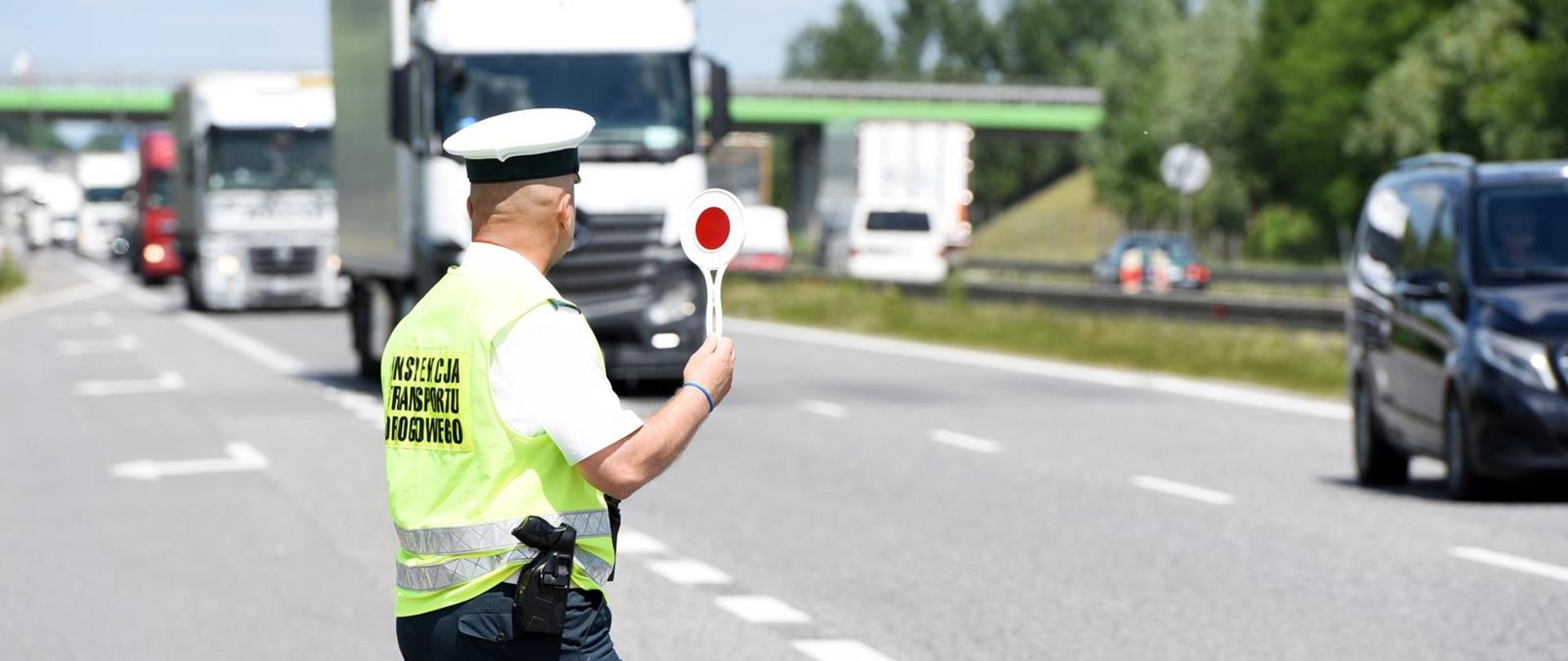 Inspektor Inspekcji Transportu Drogowego przy dwupasmowej drodze, z tarczą do zatrzymywania pojazdów w ręku. Sygnalizuje kierowcy ciężarówki zatrzymanie do kontroli. W tle pojazdy ciężarowe jadące drogą.