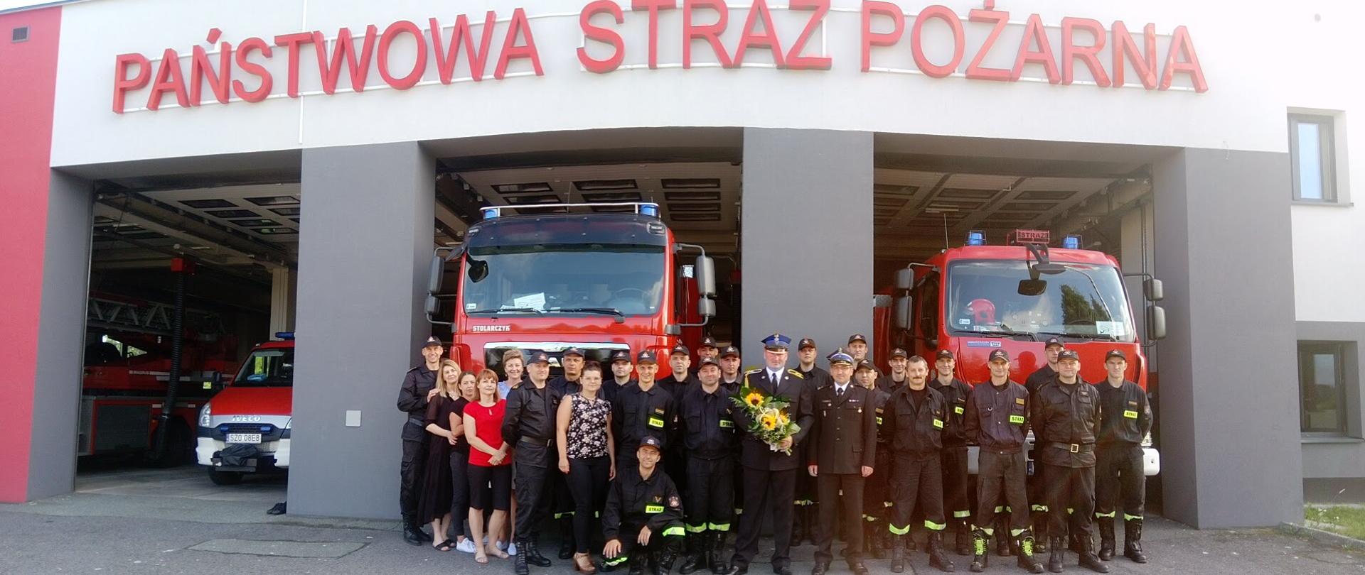Strażacy i pracownicy cywilni Komendy stojący na tle wozów strażackich. W środku Rafał Warmusz.